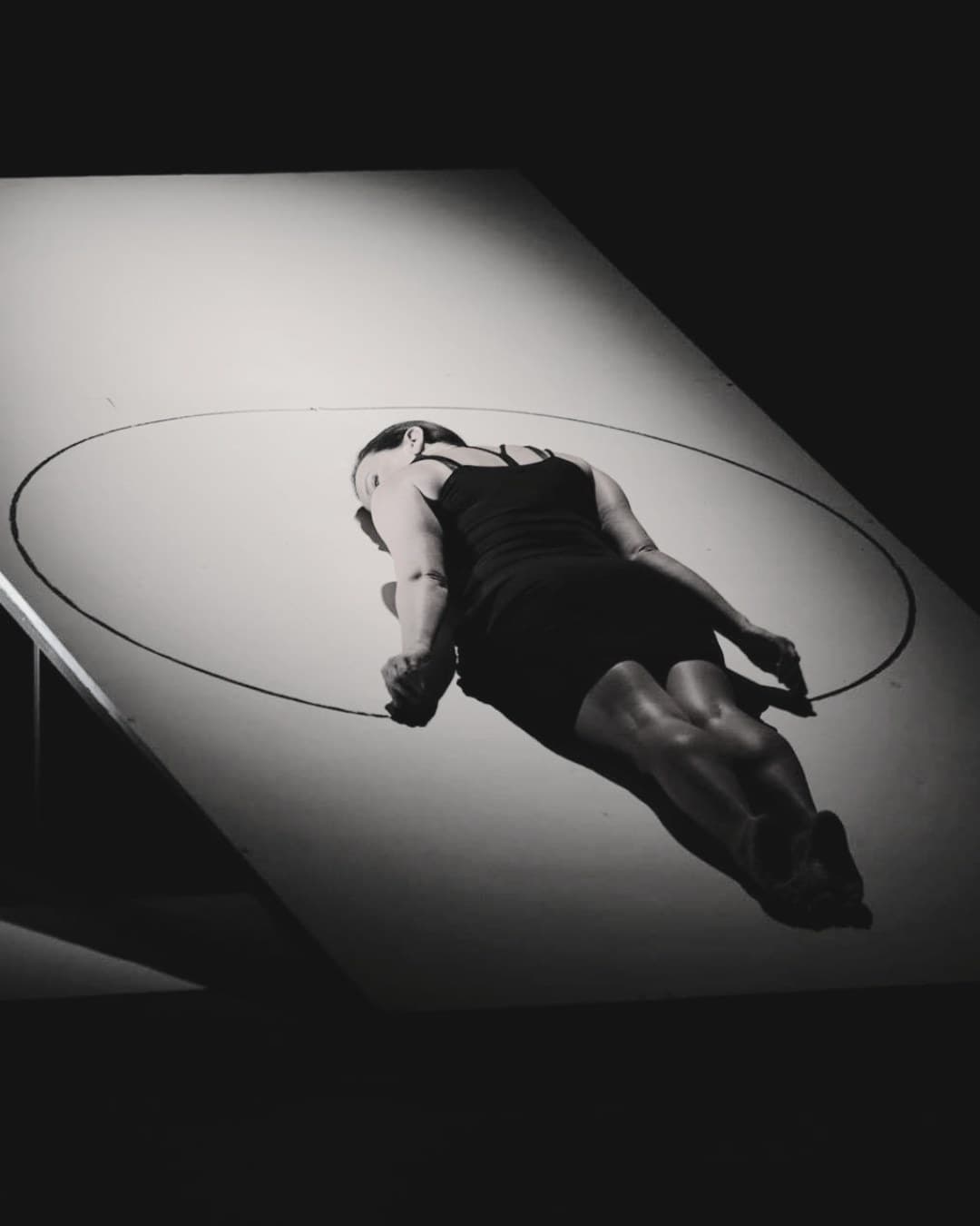 A performer lying on a brightly lit slanted stage against black, photographed in black and white.