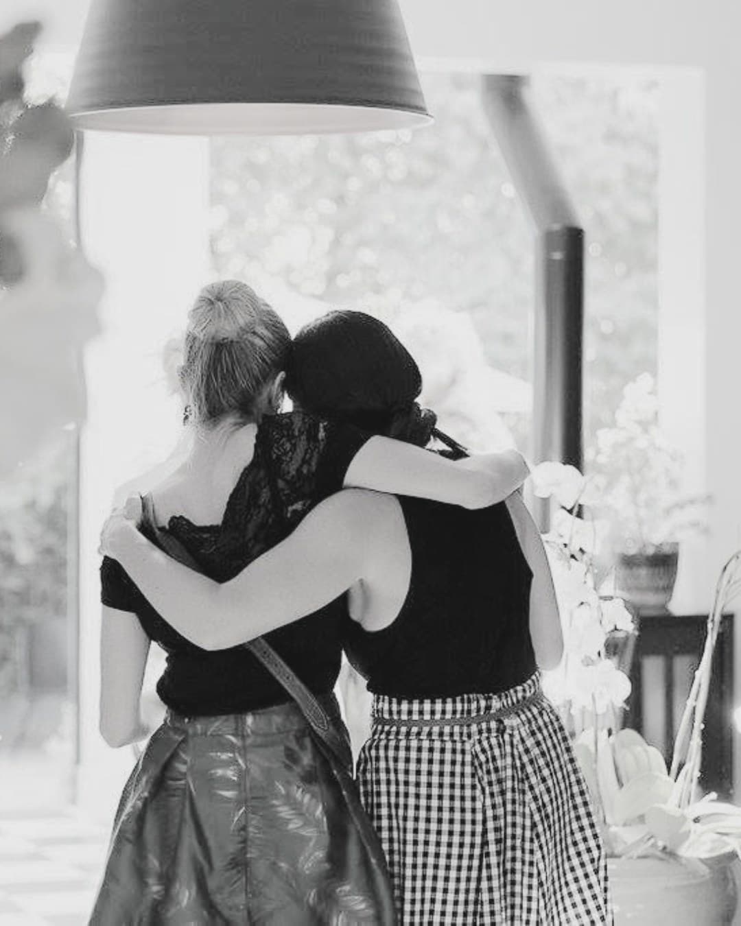 Two women standing close together with their backs to the camera, photographed in black and white.